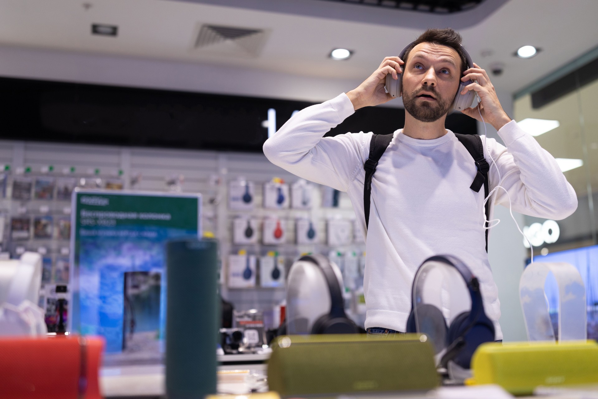 Photo of an adult handsome thinking brunet man with stubble in a white sweatshirt chooses headphones in an electronic store in a shopping mall. Shop and music concept
